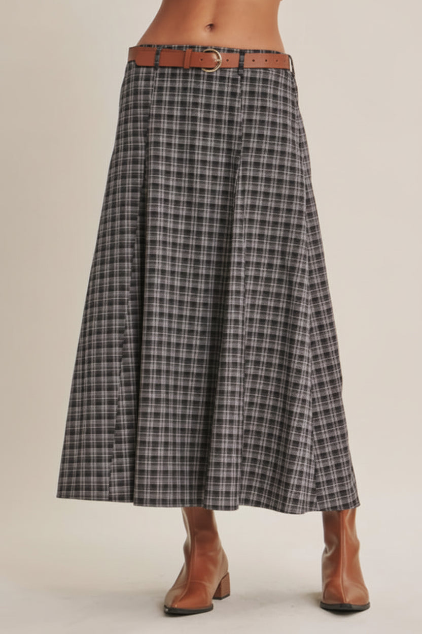 A model is wearing a high-waisted, A-line plaid skirt in gray, black, and white, styled with a brown leather belt and brown ankle boots against a solid beige background.