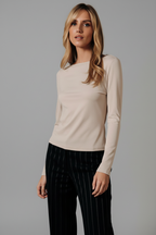A woman modeling a fitted, long-sleeve nude colored top with a boat neckline, paired with black pinstriped trousers, set against a neutral back drop