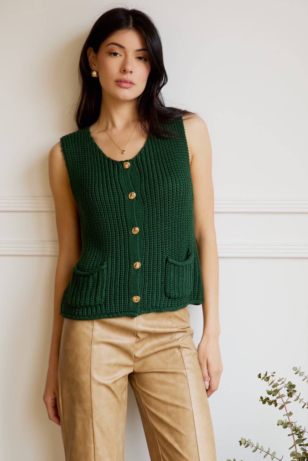 Woman wearing a green knitted vest with gold buttons against a white wall.
