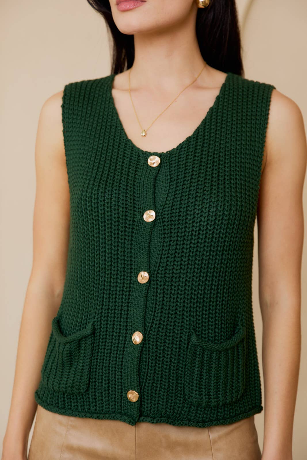 Woman wearing a green knitted vest with gold buttons against a white wall.