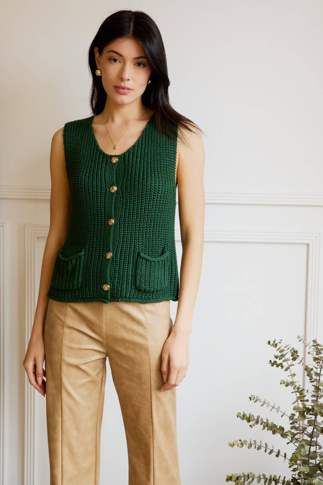 Woman wearing a green knitted vest with gold buttons and gold pants against white background.