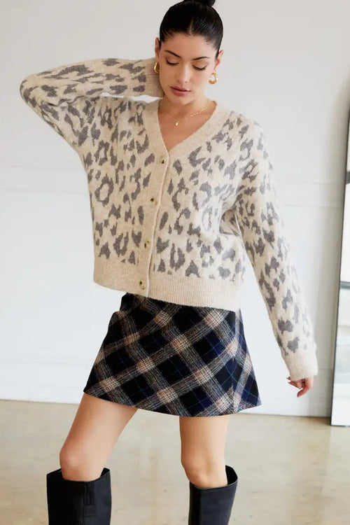 Woman wearing a leopard print cardigan, plaid skirt, and black boots in a minimal indoor setting.