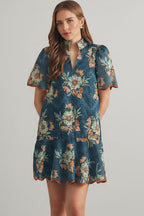 Woman wearing a teal and rust floral eyelet mini dress against a beige tiled wall.