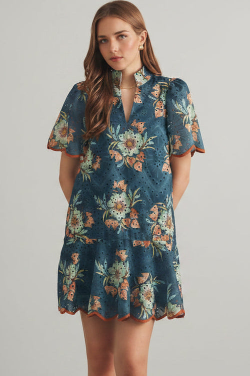 Woman wearing a teal and rust floral eyelet mini dress against a beige tiled wall.