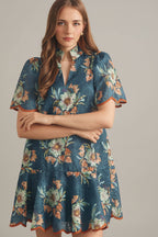 Woman wearing a teal and rust floral eyelet mini dress against a beige tiled wall.