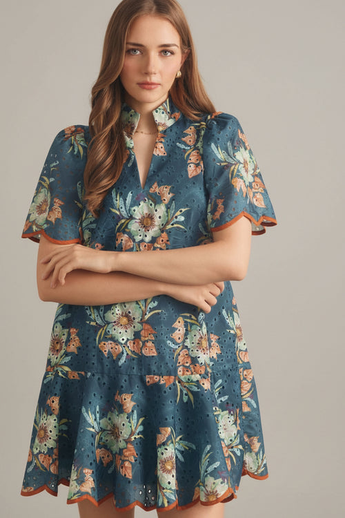Woman wearing a teal and rust floral eyelet mini dress against a beige tiled wall.