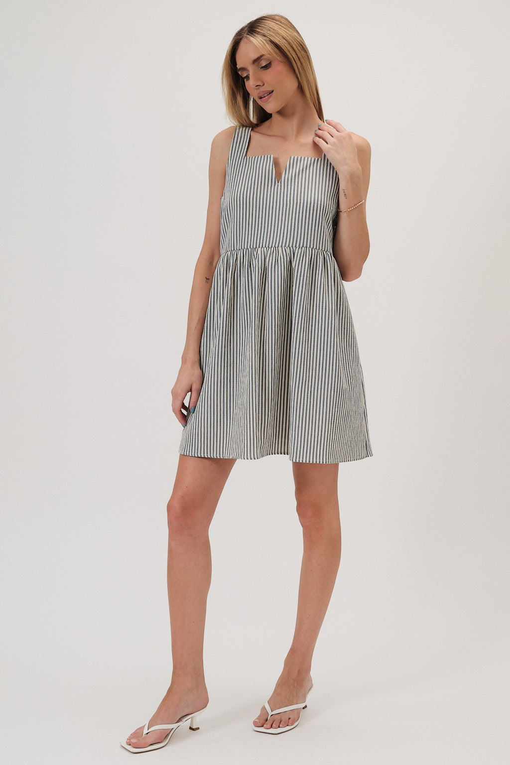 A woman wearing a sleeveless, baby doll mini dress with vertical navy and ivory stripes, paired with white heeled thong sandals, against a plain light gray studio background