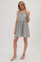 A woman wearing a sleeveless, baby doll mini dress with vertical navy and ivory stripes, paired with white heeled thong sandals, against a plain light gray studio background