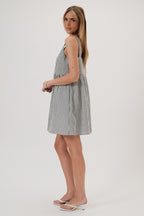A woman wearing a sleeveless, baby doll mini dress with vertical navy and ivory stripes, paired with white heeled thong sandals, against a plain light gray studio background
