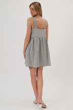 Back view of a woman wearing a sleeveless, baby doll mini dress with vertical navy and ivory stripes, paired with white heeled thong sandals, against a plain light gray studio background