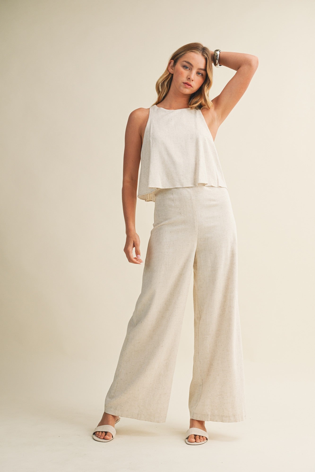 Woman wearing a cream colored sleeveless jumpsuit with overlay and wide-leg pants on a beige background