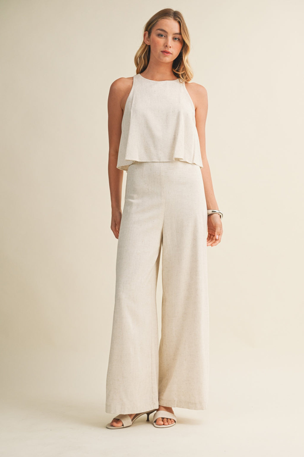 Woman wearing a cream colored sleeveless jumpsuit with overlay and wide-leg pants on a beige background