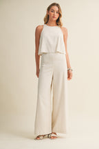 Woman wearing a cream colored sleeveless jumpsuit with overlay and wide-leg pants on a beige background
