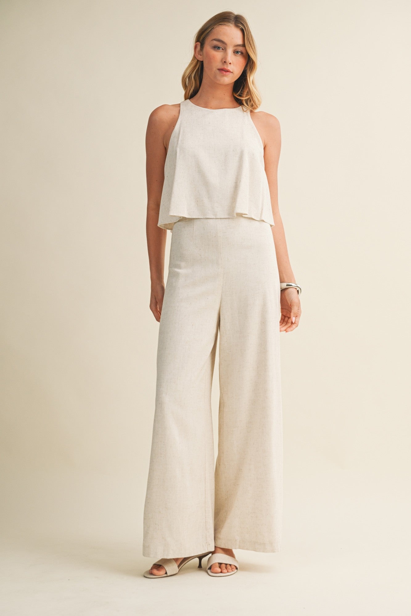 Woman wearing a cream colored sleeveless jumpsuit with overlay and wide-leg pants on a beige background