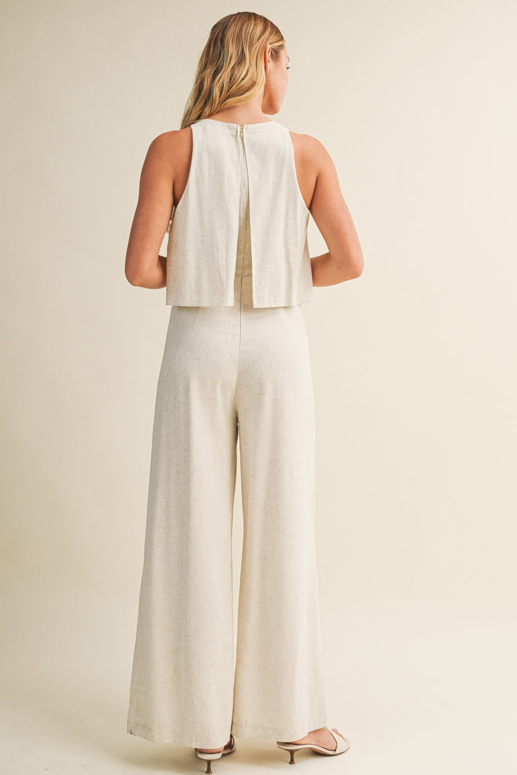 Woman wearing a cream colored sleeveless jumpsuit with overlay and wide-leg pants on a beige background
