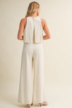 Woman wearing a cream colored sleeveless jumpsuit with overlay and wide-leg pants on a beige background