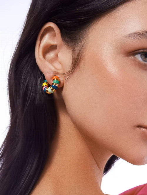 A close-up of a model's ear wearing a small hoop earring decorated with colorful enamel-like beads in orange, yellow, green, blue, and white.