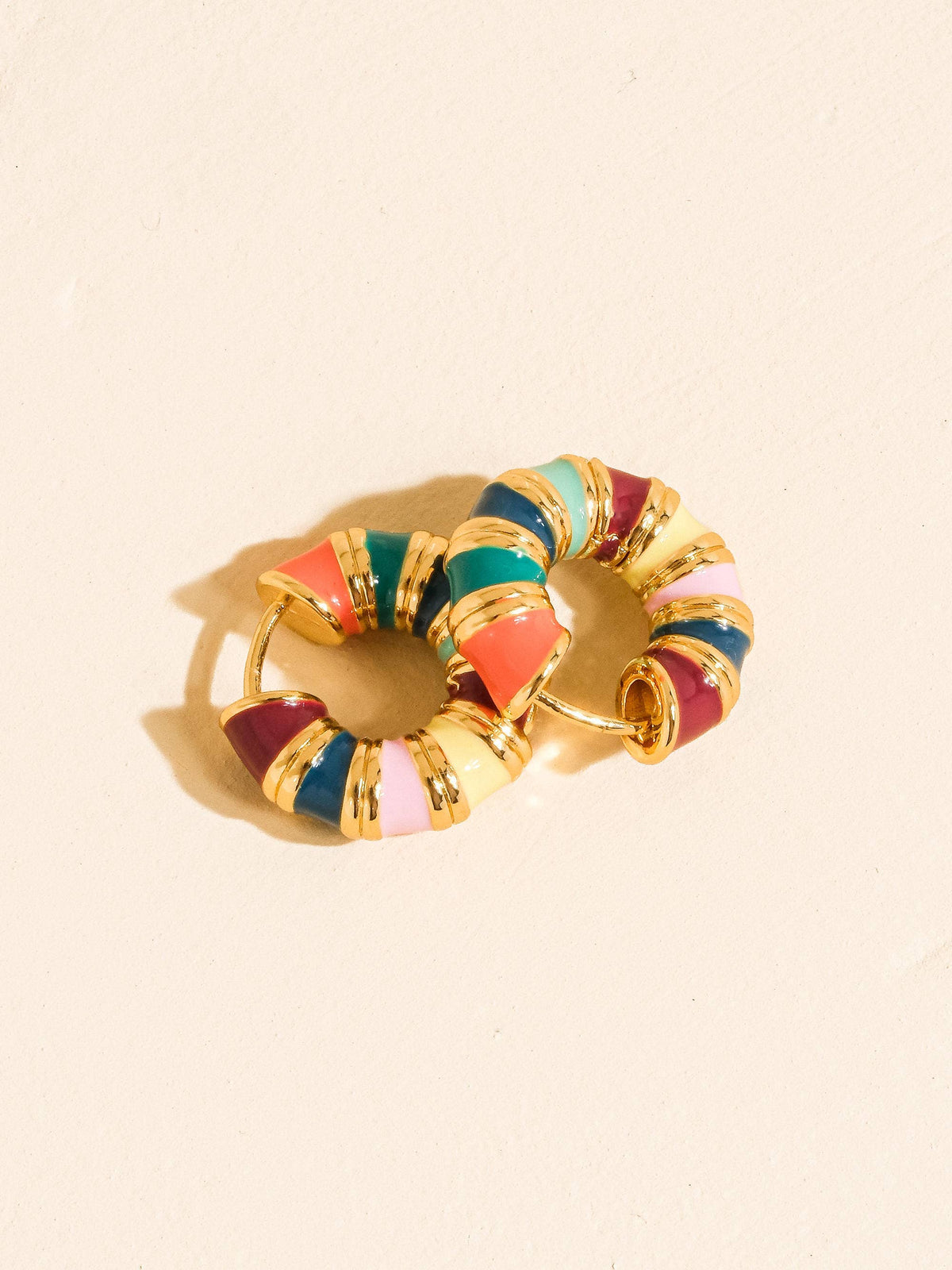 A gold-tone hoop earring with segmented, colorful enamel inlays in shades of coral, teal, burgundy, navy, and pale pink, photographed on a warm cream background.
