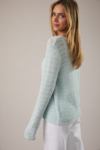 Woman wearing open weave button pale aqua pointelle knit cardigan over white tank and white jeans on a neutral background