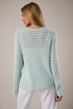 Woman wearing open weave button pale aqua pointelle knit cardigan over white tank and white jeans on a neutral background