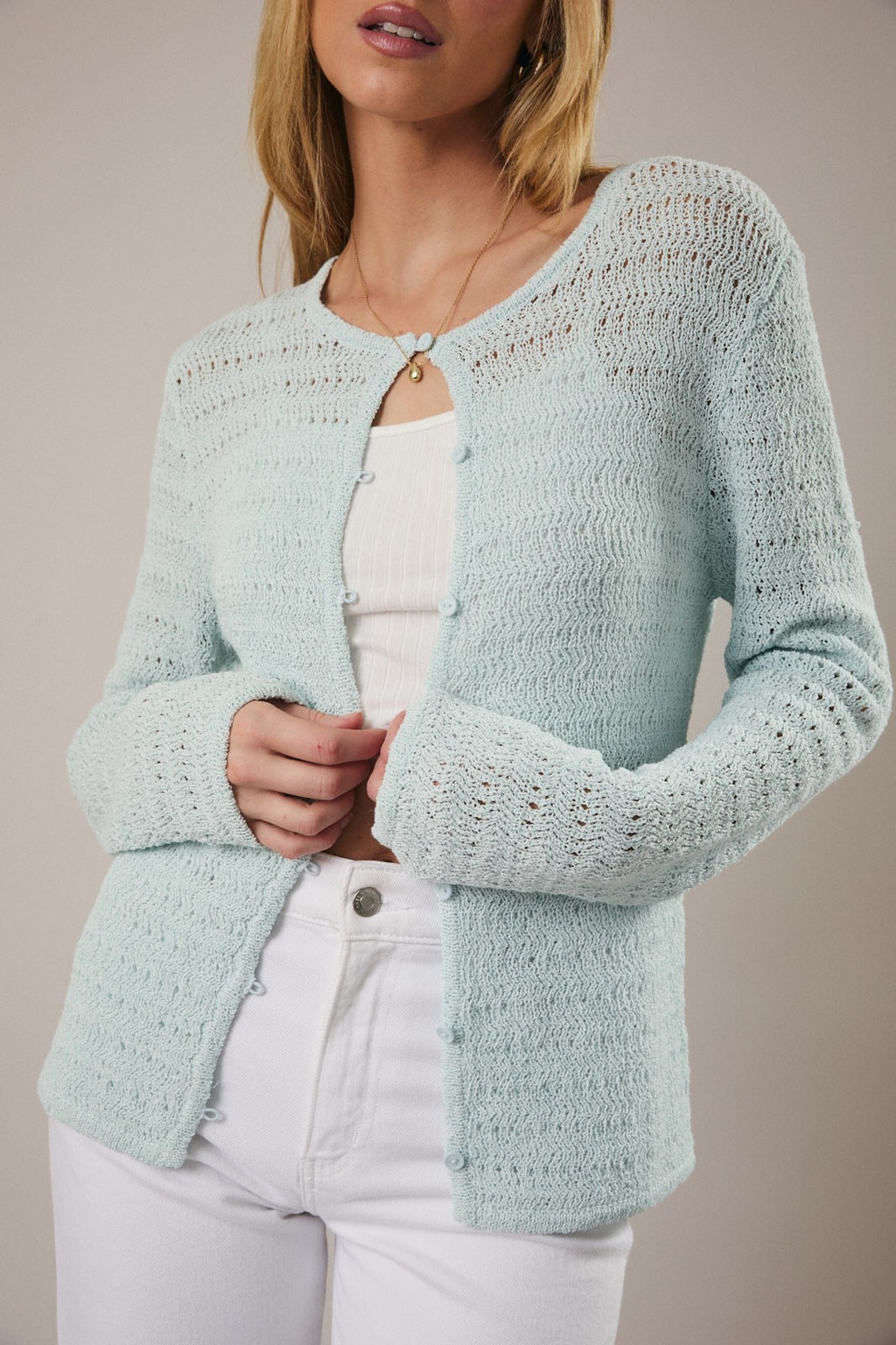 Woman wearing open weave button pale aqua pointelle knit cardigan over white tank and white jeans on a neutral background