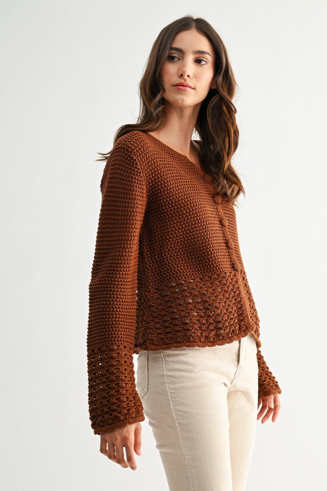 Woman wearing a chocolate brown knitted sweater with crochet detailing and white pants on a light background