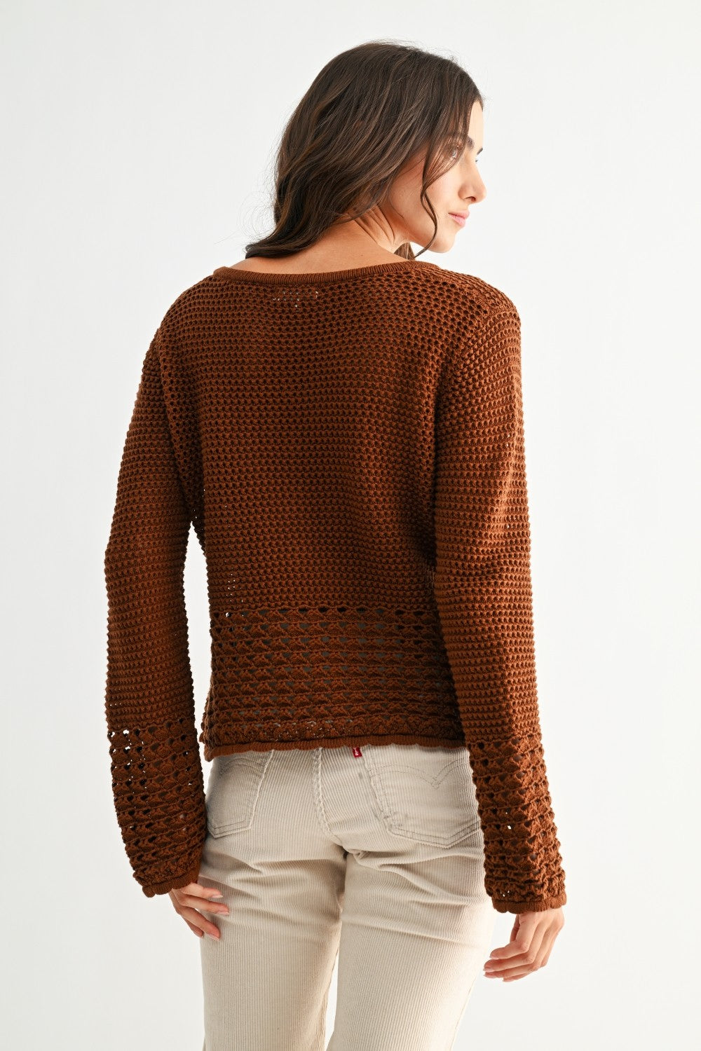 Woman wearing a chocolate brown knitted sweater with crochet detailing and light-colored pants on a white background