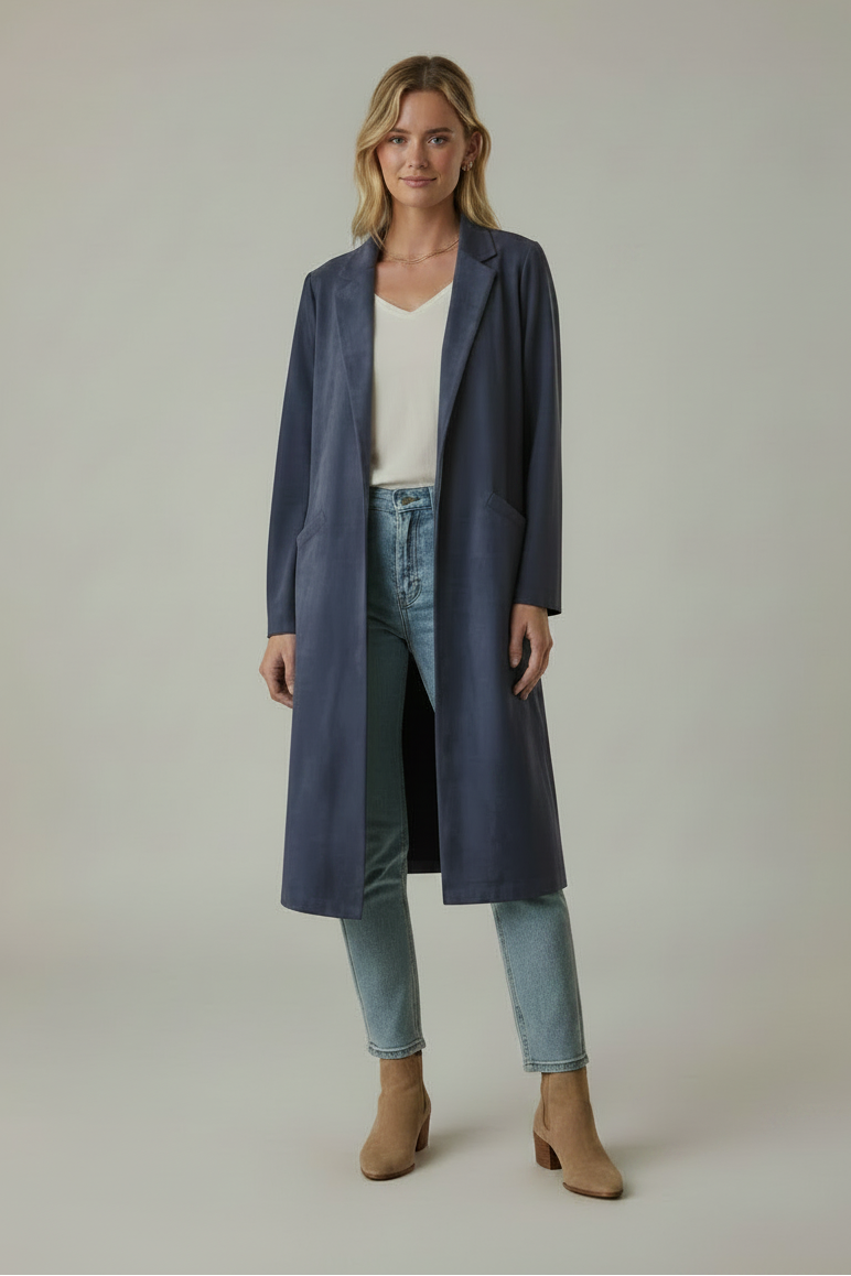 A woman wearing a long navy faux suede coat over a white top and light jeans against a grey background