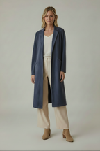 A woman stands against a plain light gray studio background wearing a long navy blue coat over a cream top and cream wide-leg pants, with tan ankle boots.