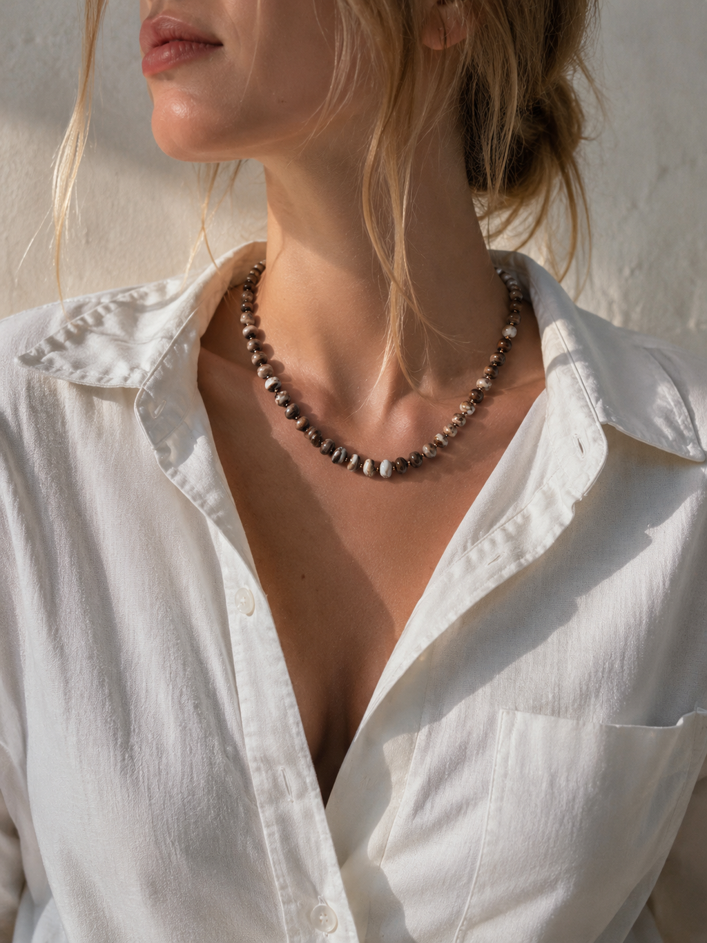 A close-up of a woman wearing a beaded necklace made of alternating multi brown and seed beads, styled with a white button-down shirt.