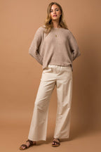 Woman wearing a taupe woven top and white pants against a brown background