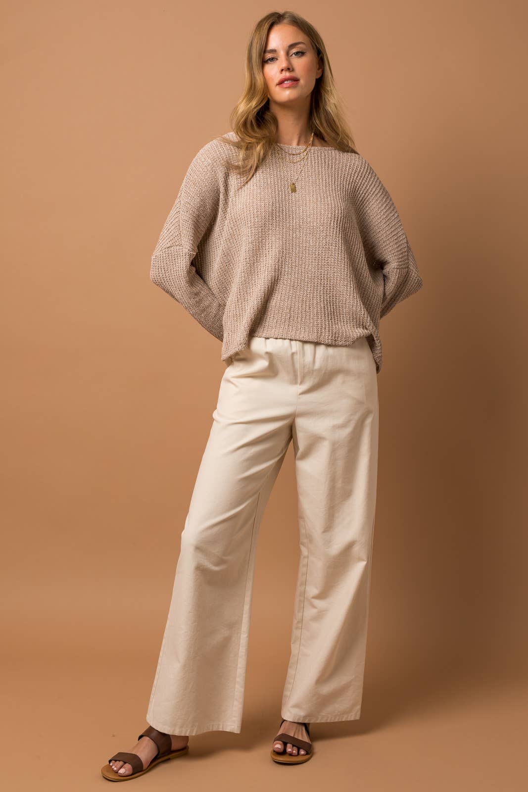 Woman wearing a taupe woven top and white pants against a brown background