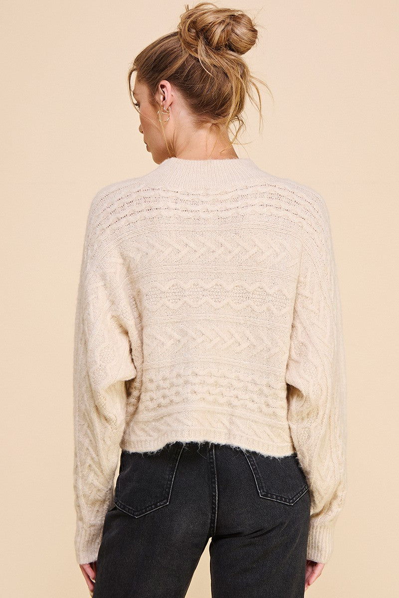 Woman showing back of cream long sleeve mock neck cable knit sweater on beight background 