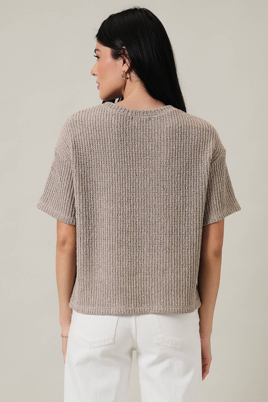 Back view of a woman wearing an oatmeal boucle knit top and white pants against a plain background