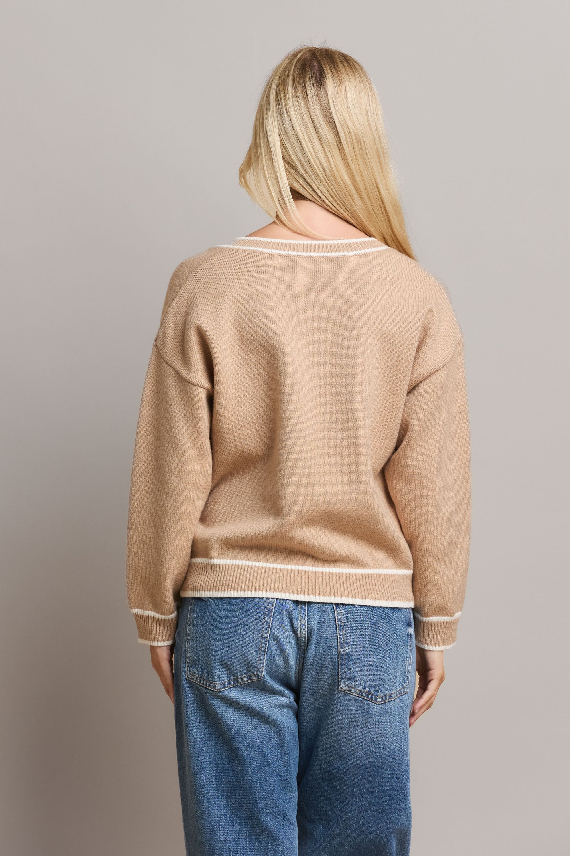 Woman wearing a tan varsity sweater with contrasting white stripe and blue jeans against a plain background