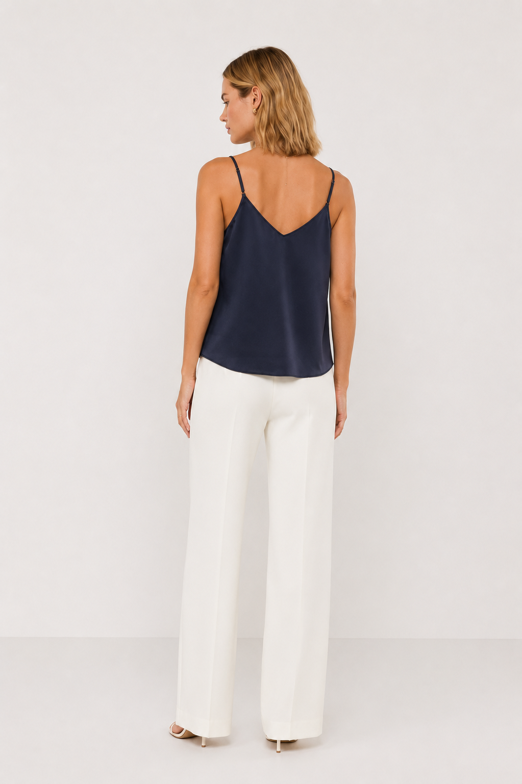 Back view of a woman wearing a navy sleeveless tank and white pants on a light gray background