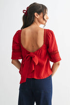 Back view of a woman wearing a scarlet lace and pleated detail top with back bow and blue jeans on a white background