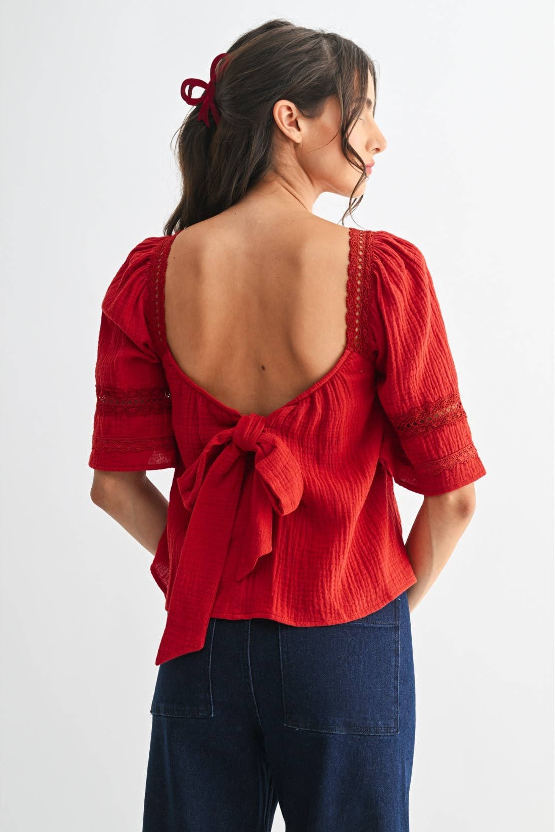 Back view of a woman wearing a scarlet lace and pleated detail top with back bow and blue jeans on a white background
