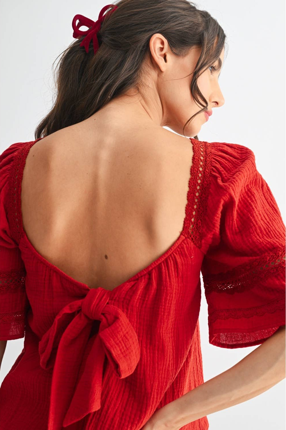 Back view of a woman wearing a scarlet lace and pleated detail top with back bow and blue jeans on a white background