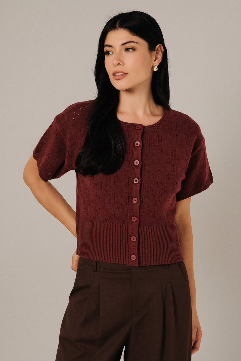 A woman modeling a short-sleeve berry  pointelle knit cardigan with a button-down front, paired with white pants, set against a soft white curtain background.