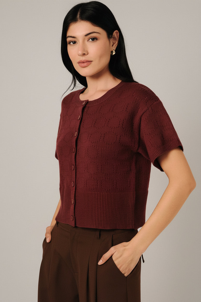 A woman modeling a short-sleeve berry  pointelle knit cardigan with a button-down front, paired with white pants, set against a soft white curtain background.
