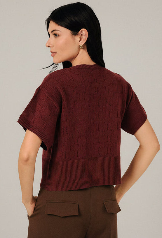 Back view of a woman modeling a short-sleeve berry  pointelle knit cardigan with a button-down front, paired with white pants, set against a soft white curtain background.