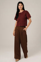 A woman modeling a short-sleeve berry  pointelle knit cardigan with a button-down front, paired with white pants, set against a soft white curtain background.
