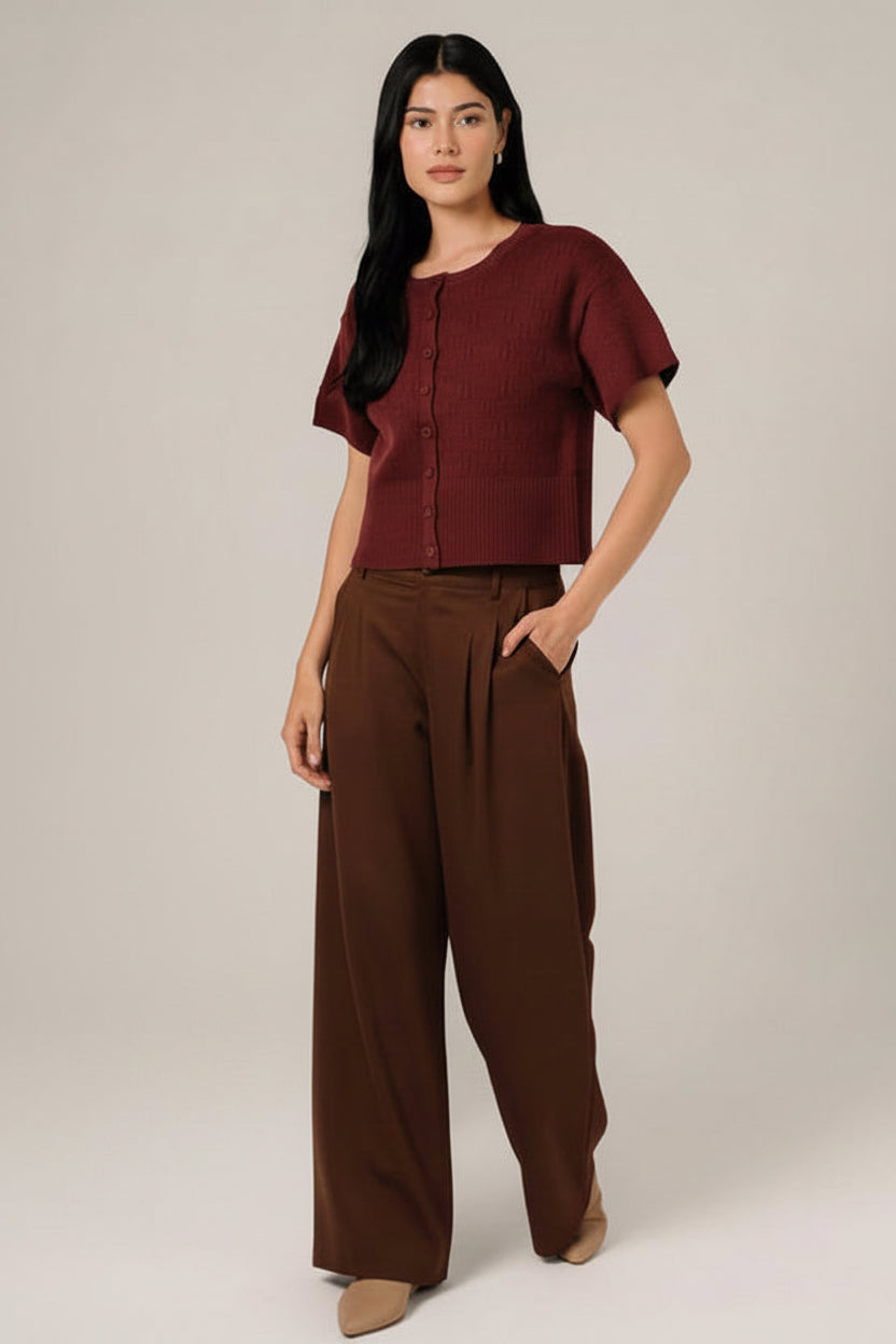 A woman modeling a short-sleeve berry  pointelle knit cardigan with a button-down front, paired with white pants, set against a soft white curtain background.