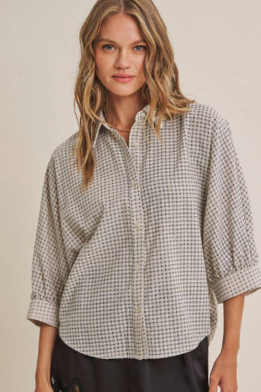 A woman modeling a three-quarter sleeve button-up shirt with navy and cream micro plaid fabric paired with black shorts, against a solid beige studio background.