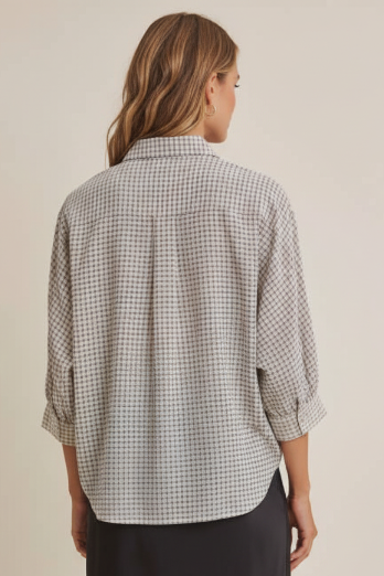 Back view of a woman modeling a three-quarter sleeve button-up shirt with navy and cream micro plaid fabric paired with black skirt, against a solid beige studio background.