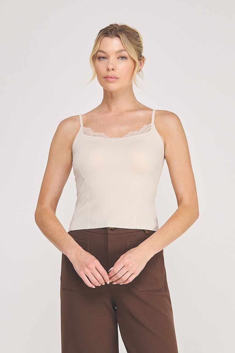 A woman modeling a light beige camisole top with delicate eyelash lace trim at the neckline, paired with brown pants, against a plain light gray studio background.