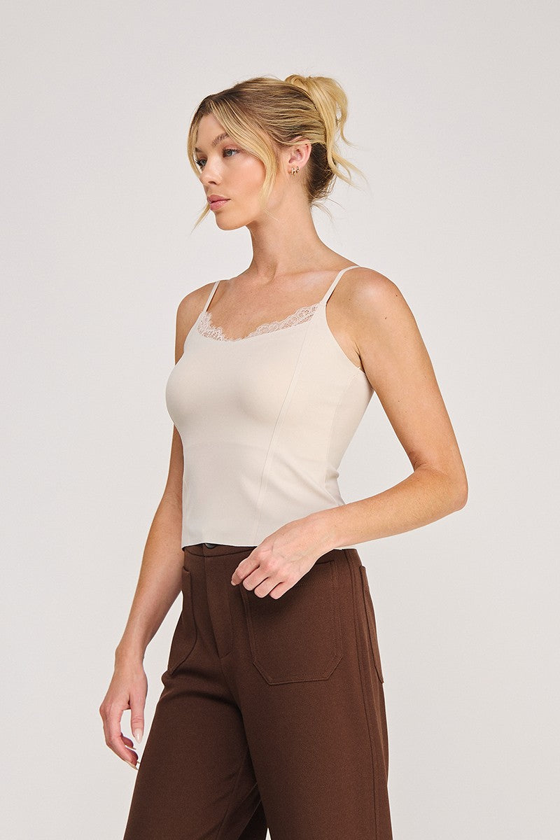 A woman modeling a light beige camisole top with delicate eyelash lace trim at the neckline, paired with brown pants, against a plain light gray studio background.