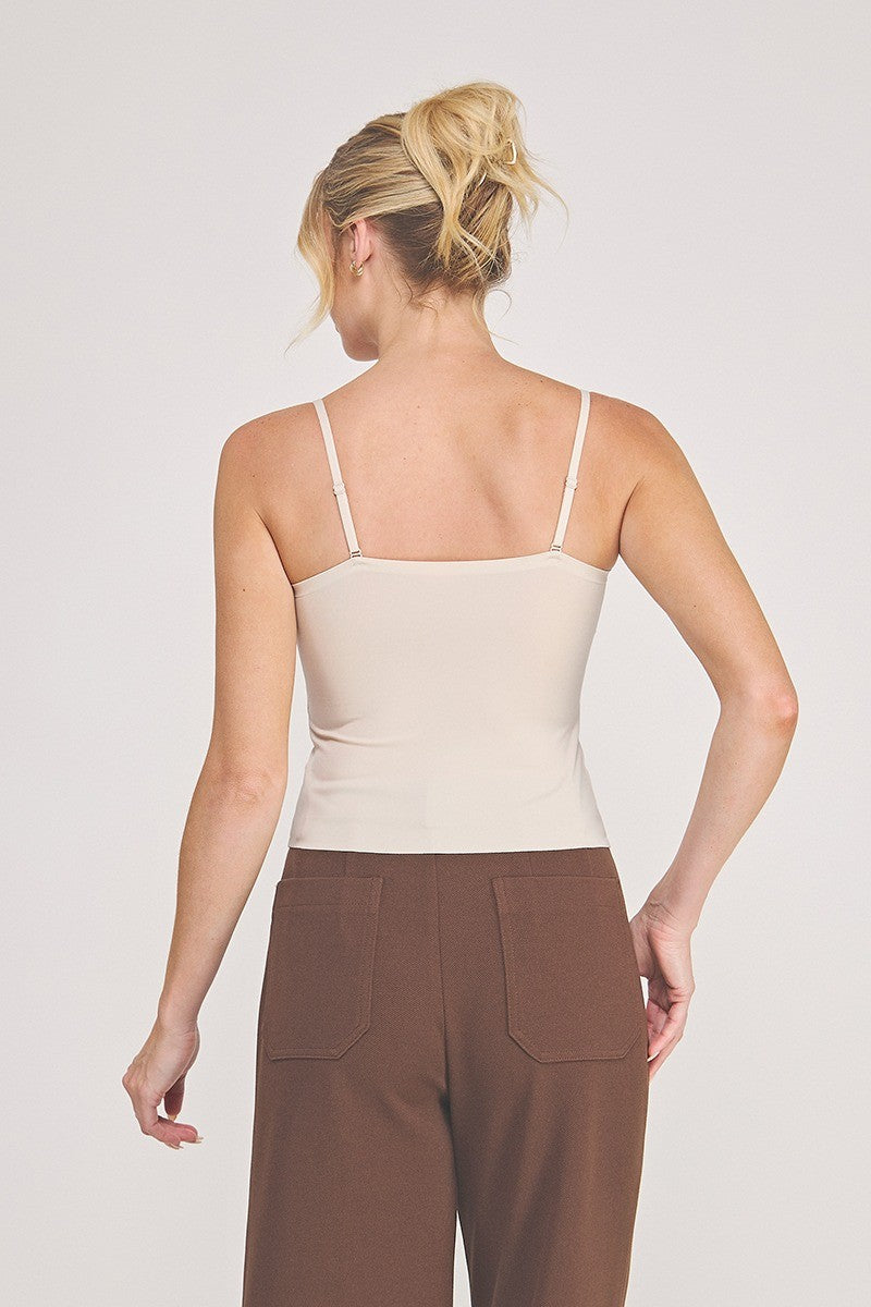 Back view of a woman modeling a light beige camisole top with delicate eyelash lace trim at the neckline, paired with brown pants, against a plain light gray studio background.