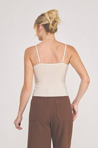 Back view of a woman modeling a light beige camisole top with delicate eyelash lace trim at the neckline, paired with brown pants, against a plain light gray studio background.